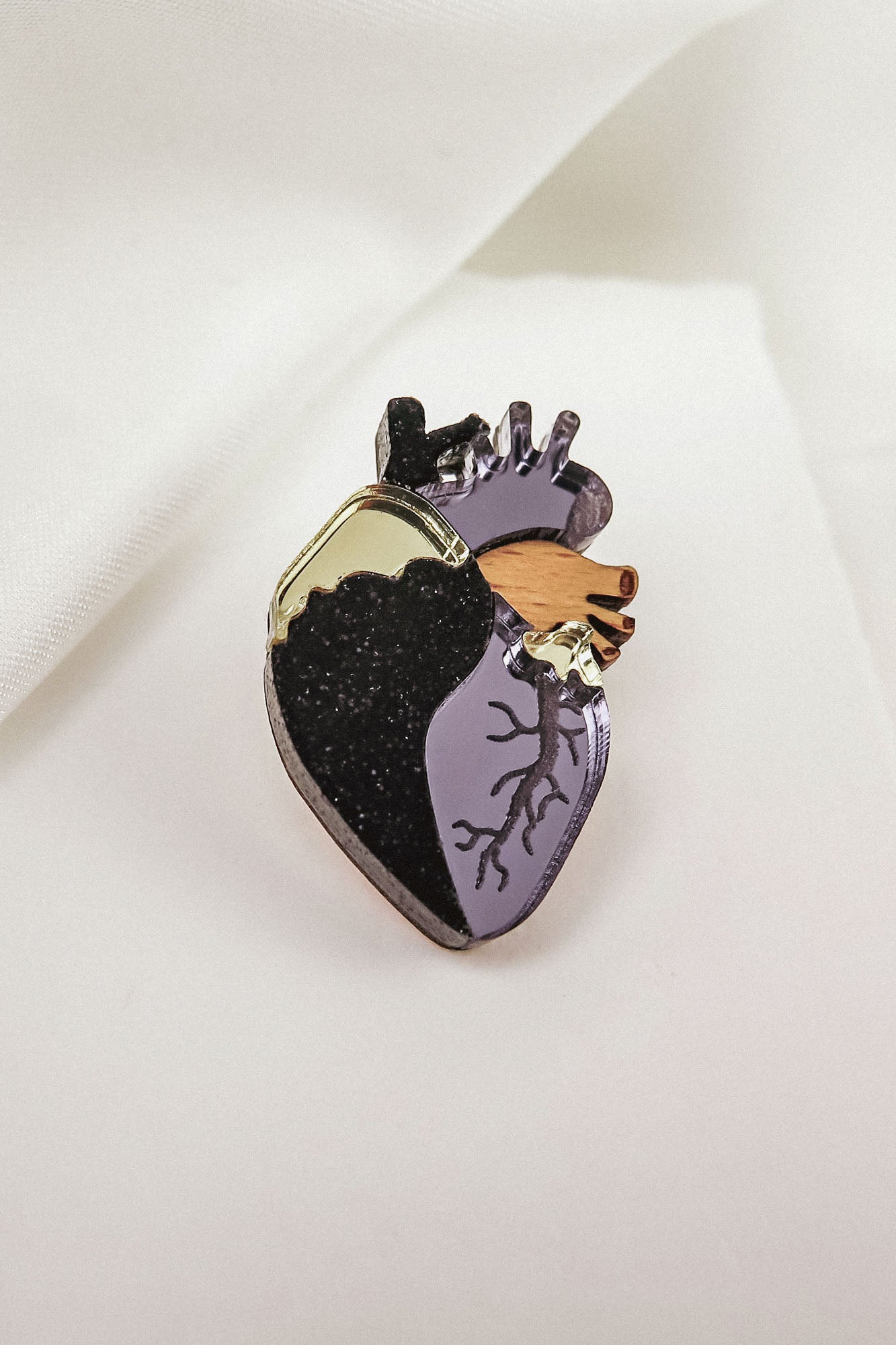 A handcrafted Anatomical Heart Pin, this decorative enamel statement piece features black, gold, and purple sections with detailed veins, beautifully displayed on a white fabric background.