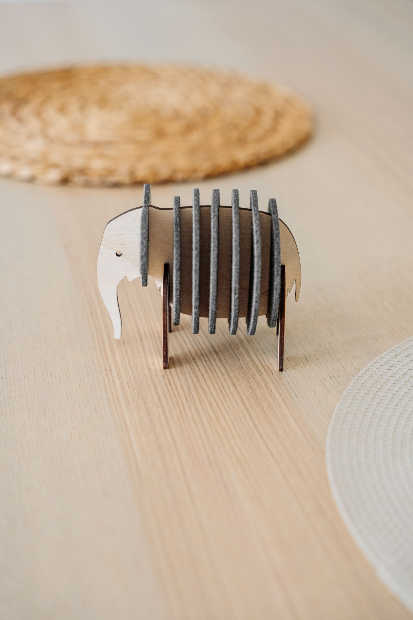 A small, elephant-shaped coaster holder made of wood and dark felt stands on a light wooden table, showcasing witty design for furniture protection, surrounded by round woven placemats.