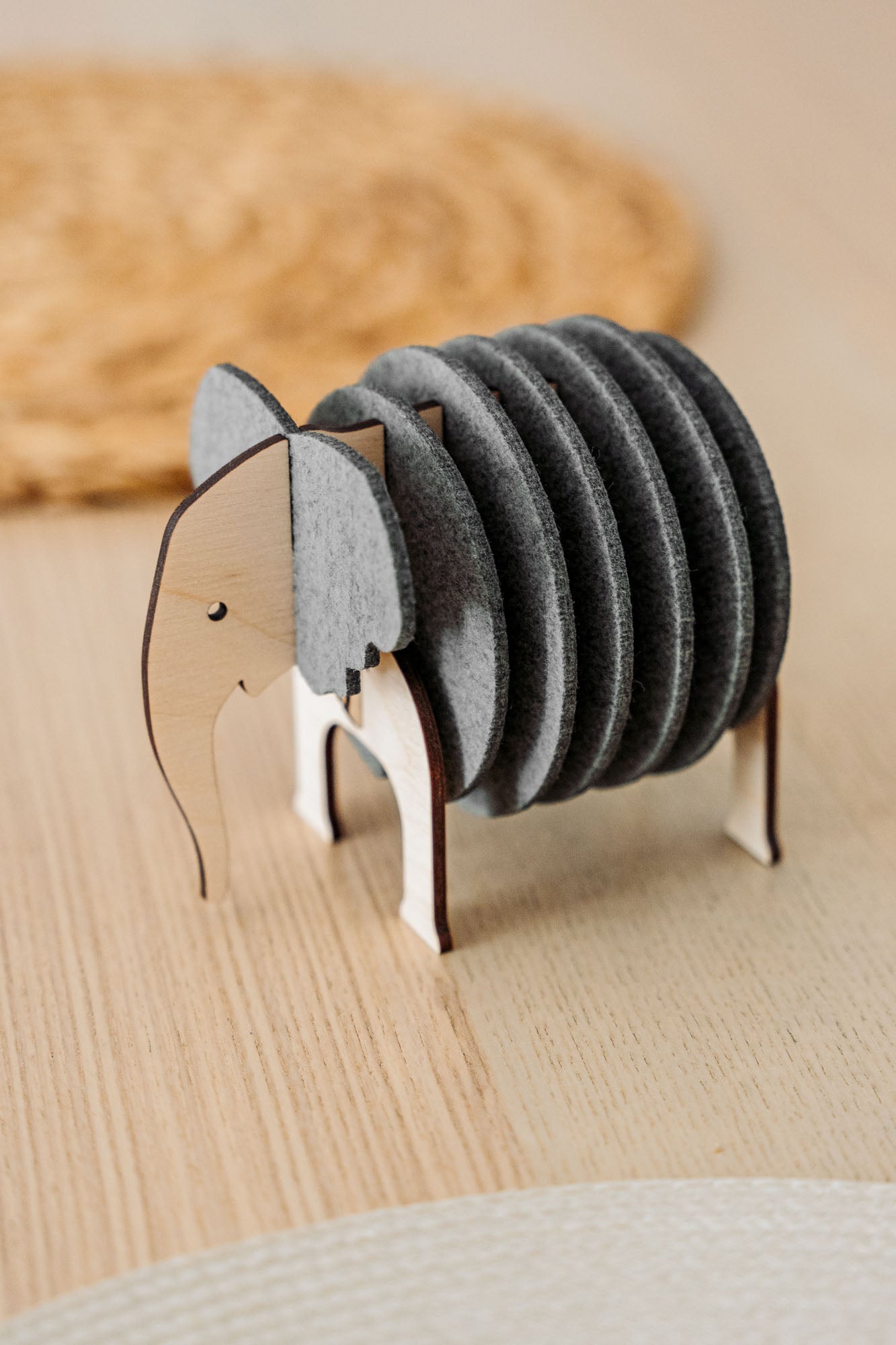 A wooden and felt Elephant Coasters holder with witty design stands on a wooden table, offering both charm and furniture protection, while a round woven placemat is blurred in the background.