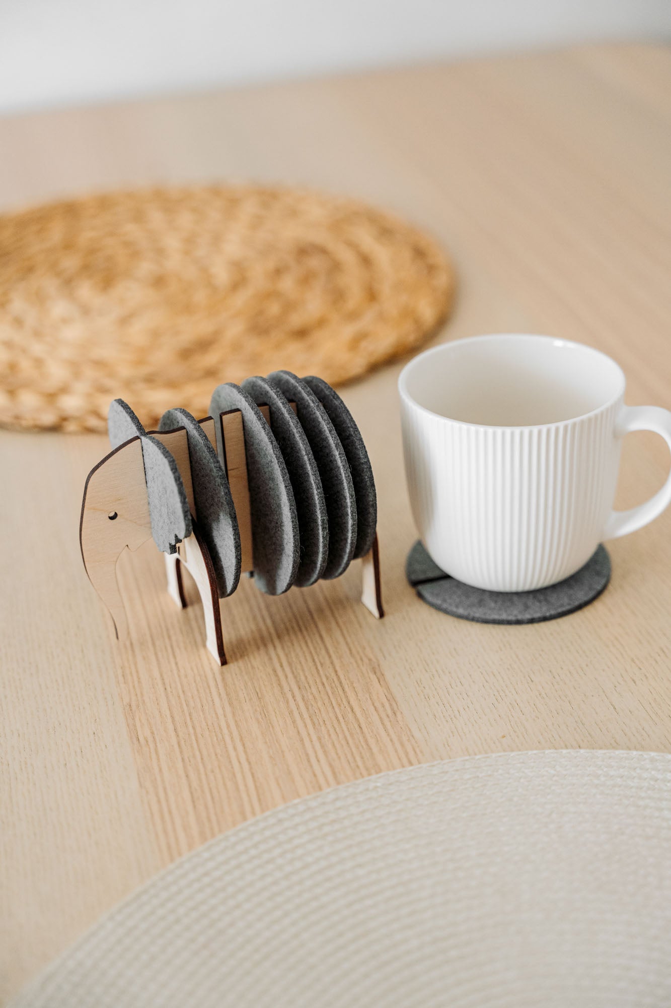 A set of round Elephant Coasters with witty design sits in a wooden elephant-shaped holder on a table, offering furniture protection while a white mug rests on one coaster.