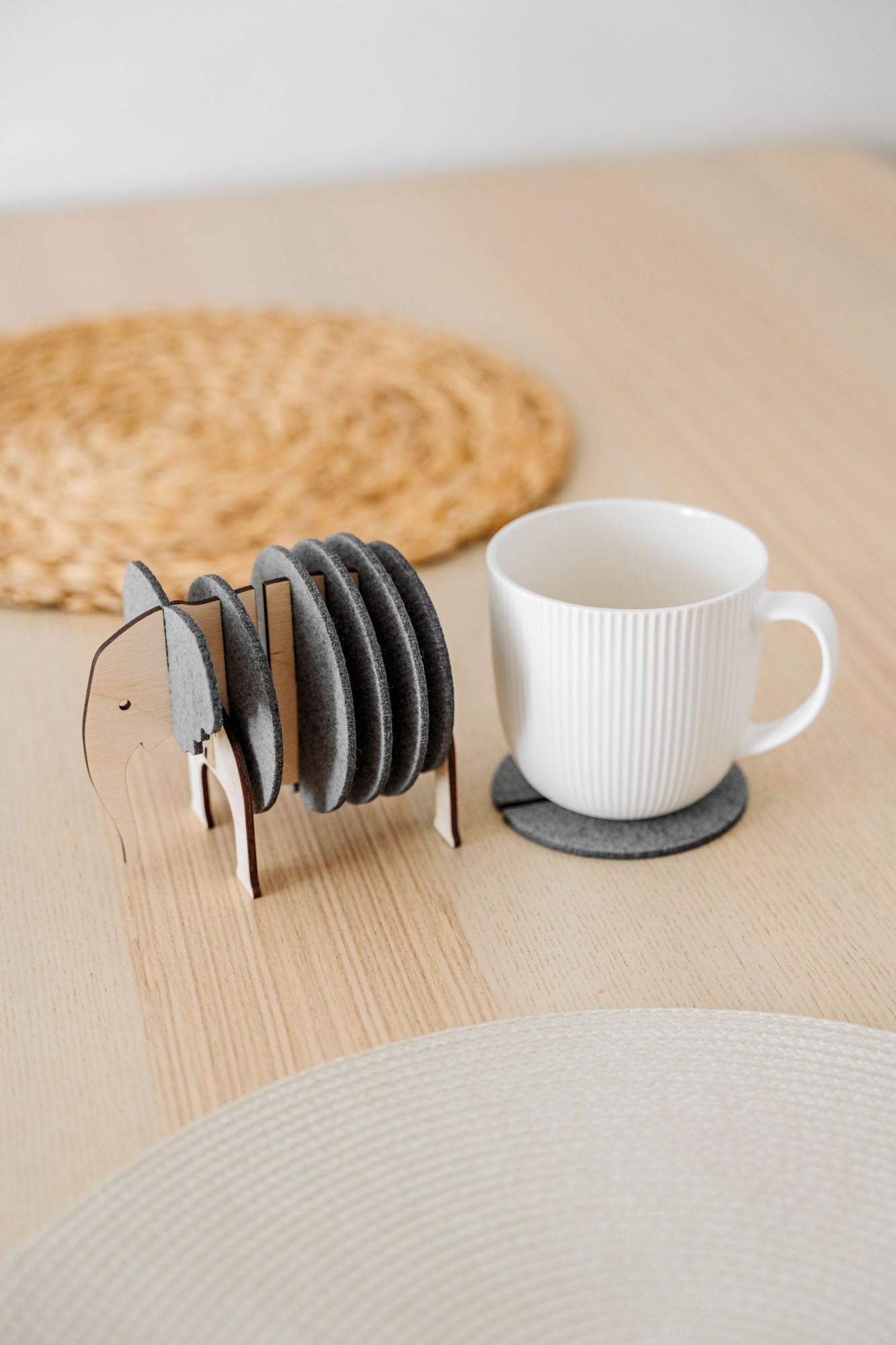 A white ribbed coffee cup sits on a round gray coaster. Beside it, Elephant Coasters with witty design are stacked in a wooden elephant-shaped holder, providing both style and furniture protection on the light wooden table. A woven placemat is in the background.