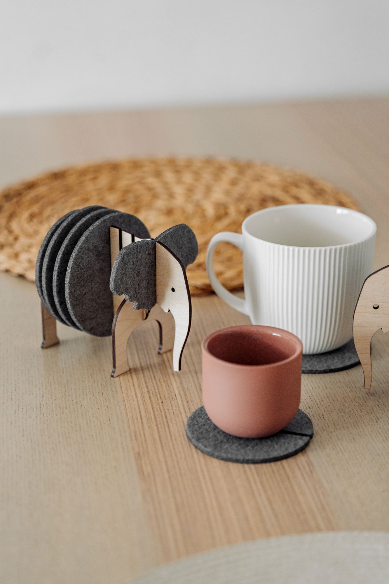 A set of Elephant Coasters with witty design offers stylish furniture protection, nestled in a wooden elephant-shaped holder on a wooden table beside a pink ceramic cup, white mug, and woven placemat.