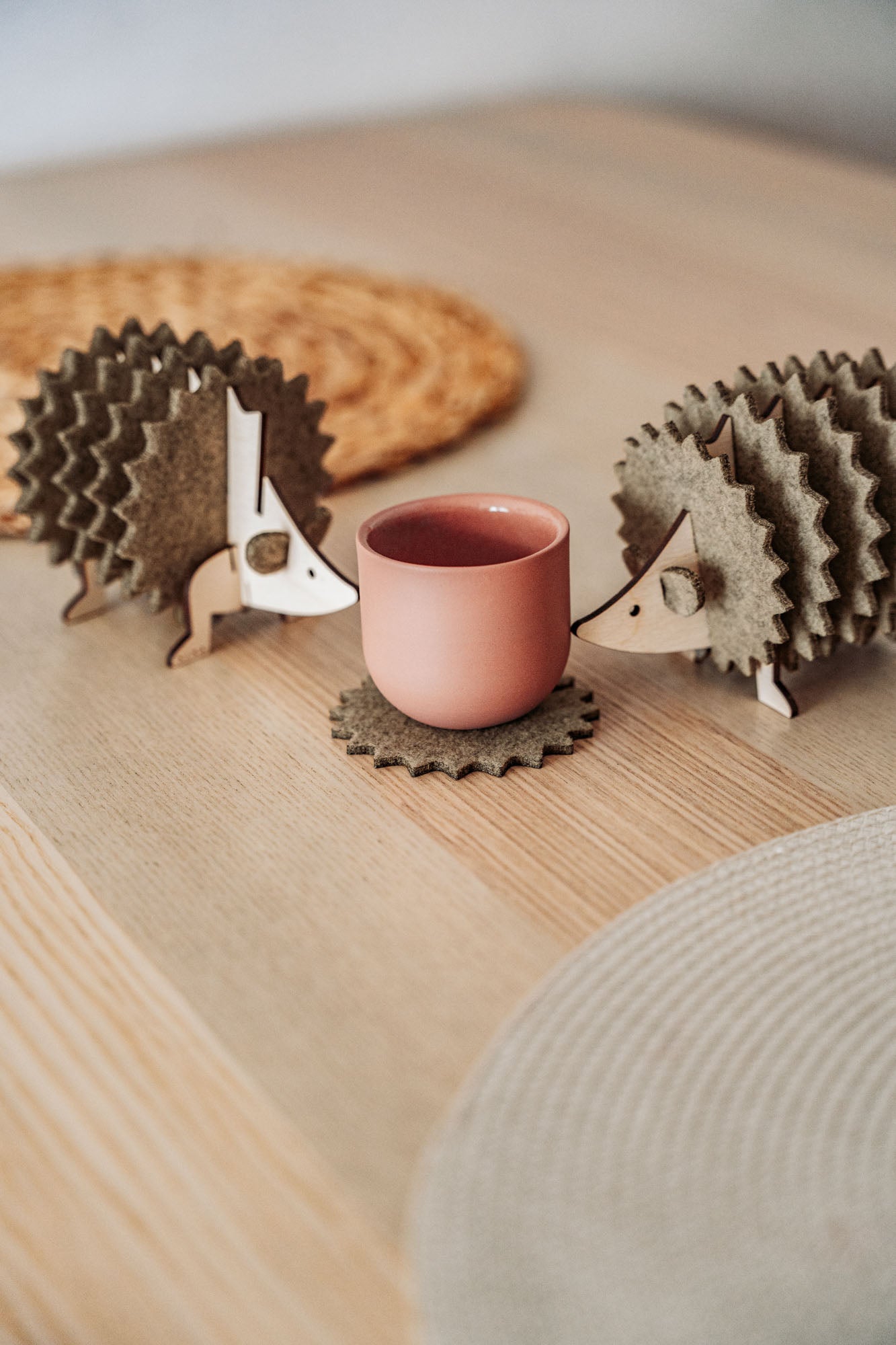 Two wooden hedgehog-shaped felt coasters hold a round, pink cup on a wooden table with woven placemats in the background—a charming touch of home decor.