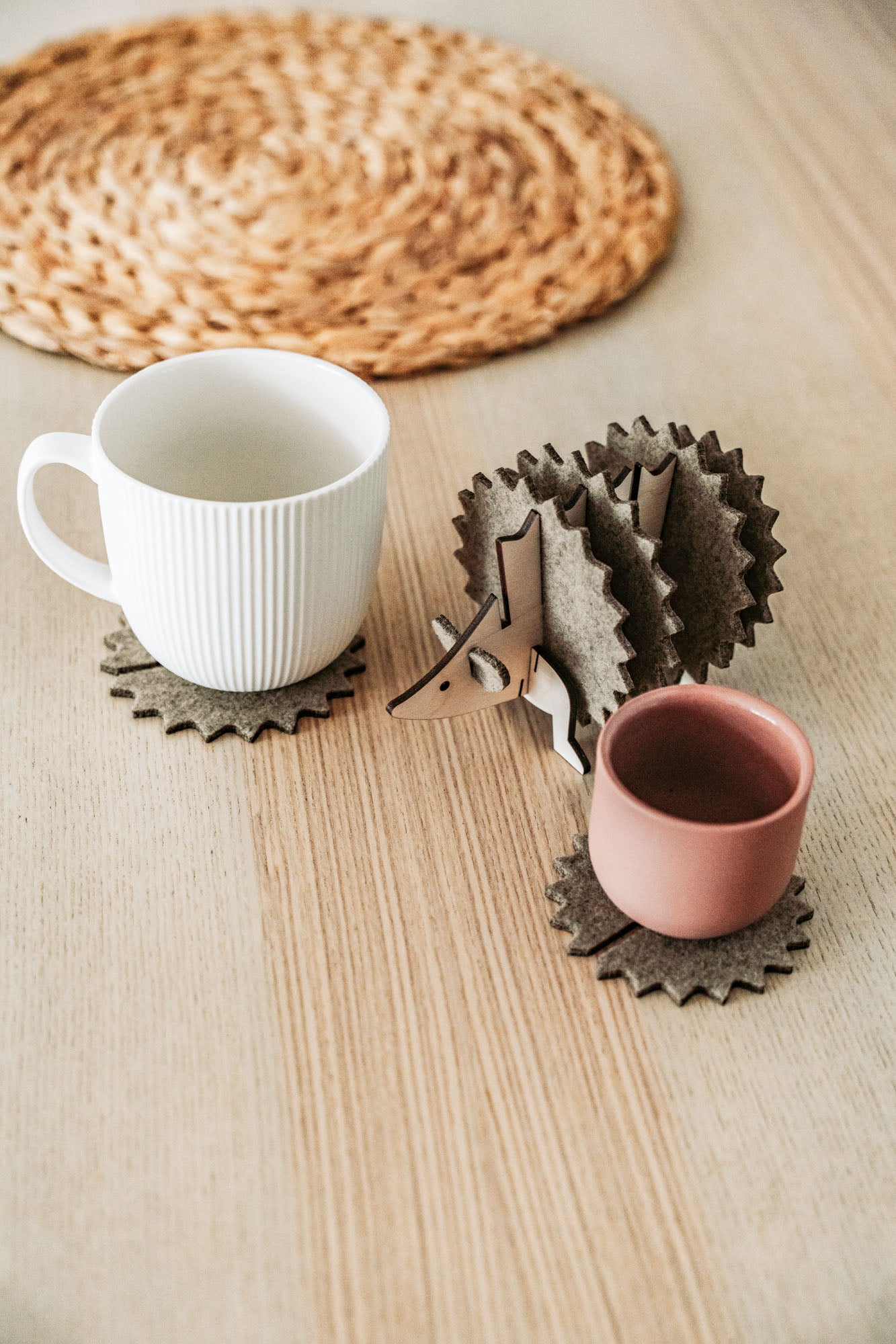 A white mug and a pink cup sit on spiky felt coasters shaped like a hedgehog, adding playful home decor flair to a light wooden table with a woven placemat in the background.