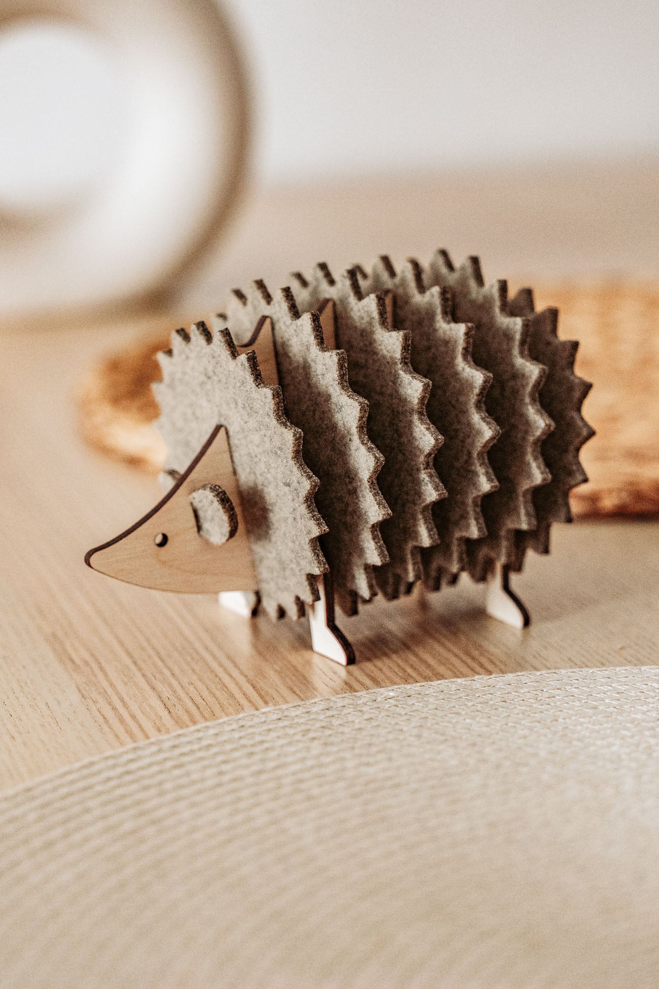 A charming wooden hedgehog coasters holder, perfect for home decor, sits on a light table with a woven placemat and blurred accents in the background.