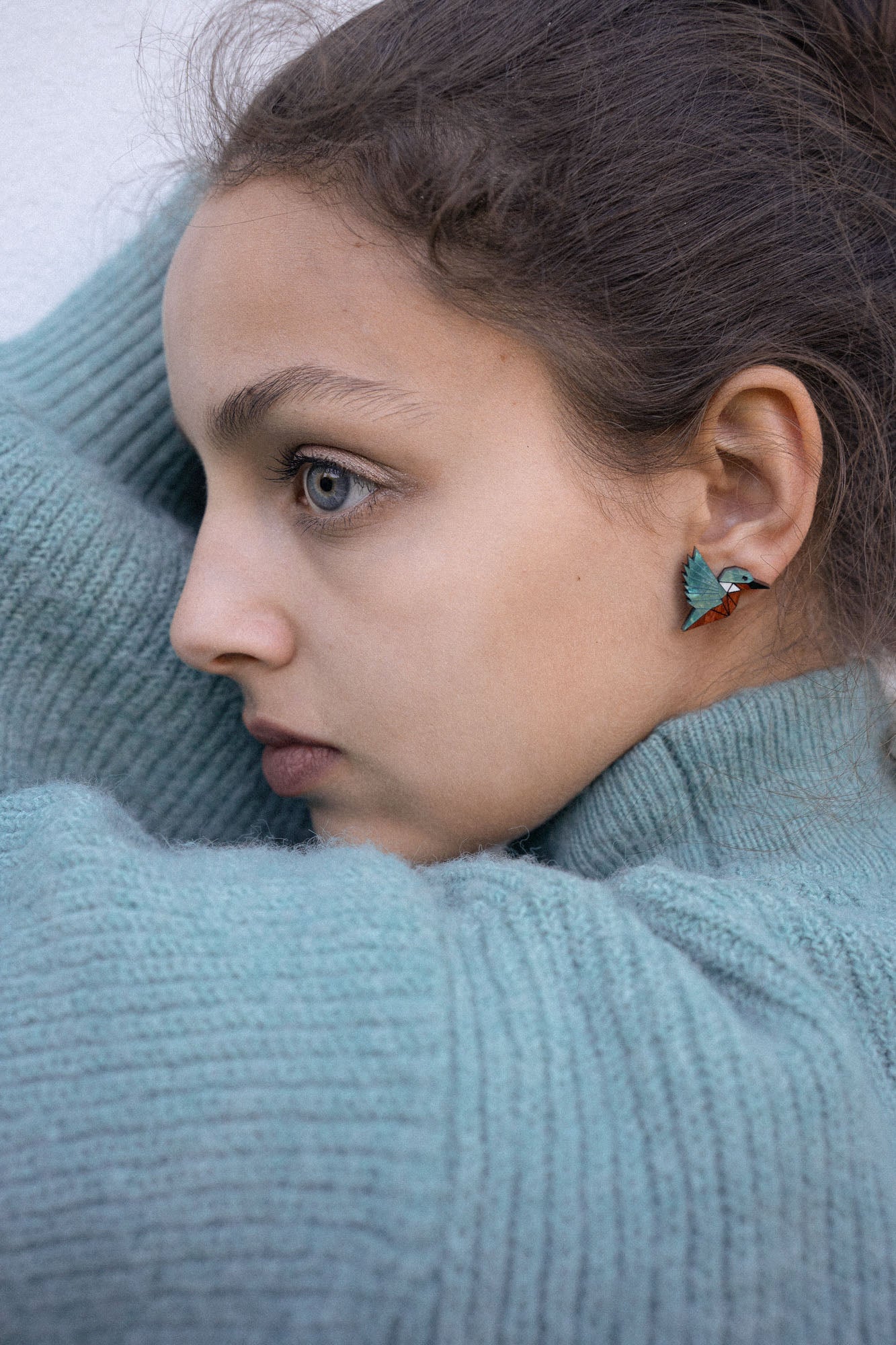 A young person wearing a teal knit sweater looks to the side, showing a Kingfisher Stud Earring. Their hair is pulled back, and their expression appears calm and thoughtful, embracing an eco-friendly, sustainably made accessory.