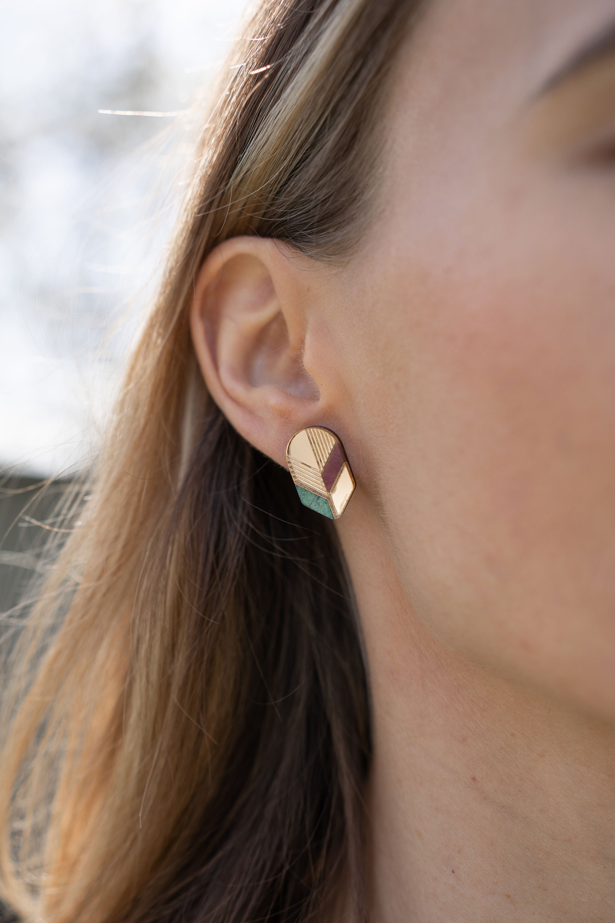 Close-up of a womans ear wearing a geometric, gold-toned stud earring with pastel accents. This piece of minimalist jewelry stands out against the softly blurred background, highlighting its unique handmade design and part of her hair and face.
