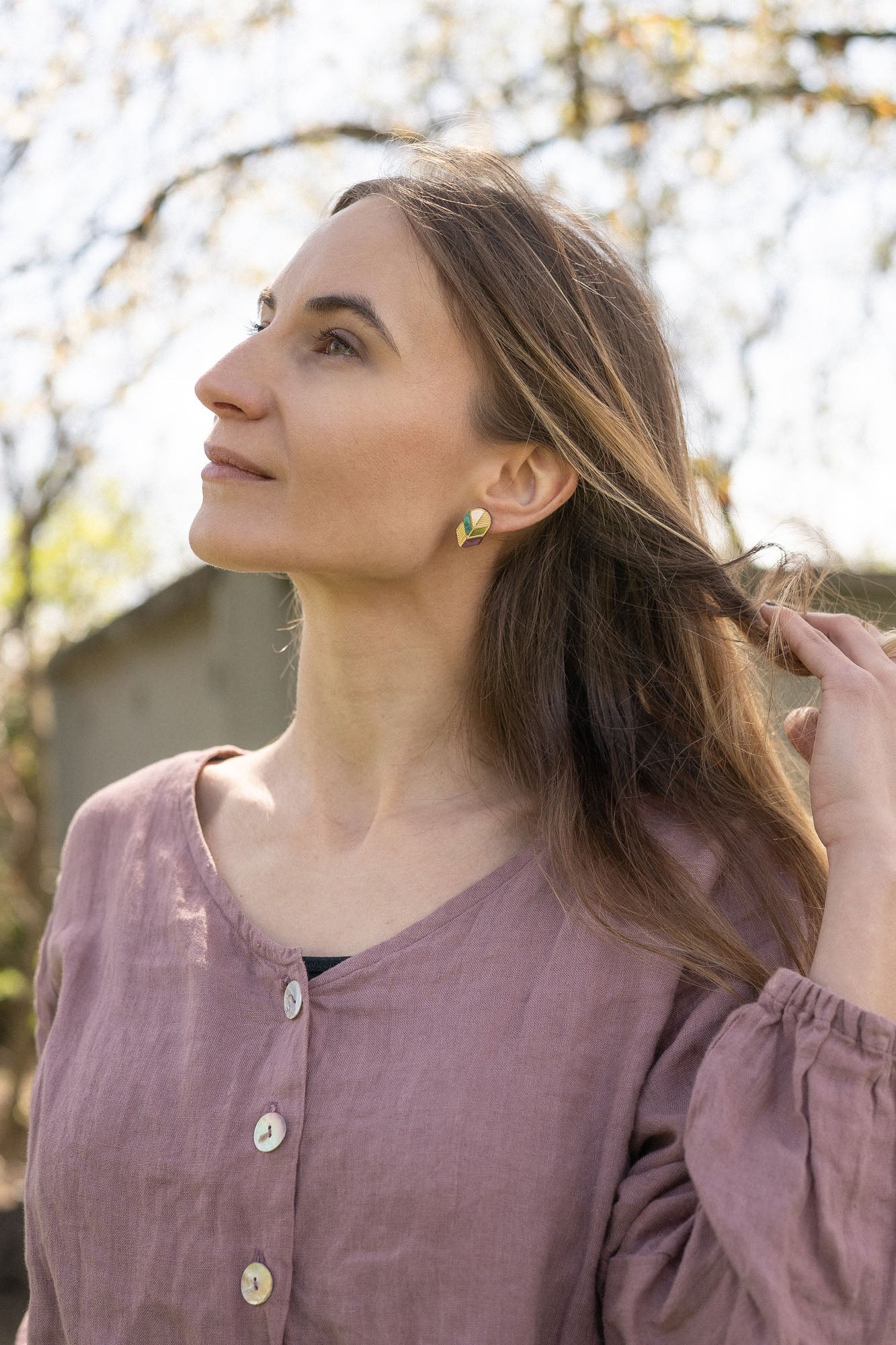 A woman with long brown hair wearing a mauve blouse and minimalist jewelry—heart-shaped earrings—looks upward while gently touching her hair; soft natural light and a blurred outdoor background.