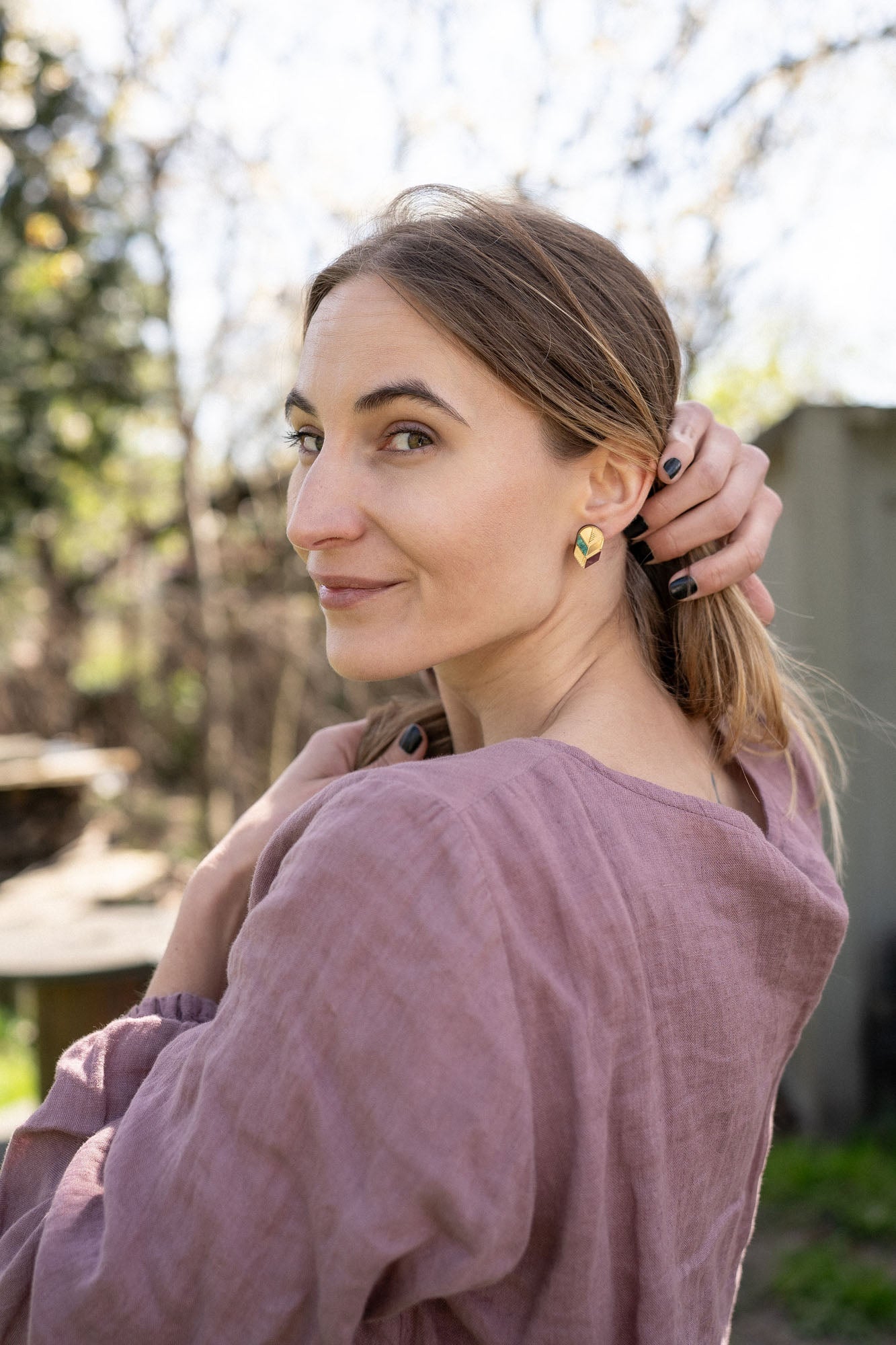 A woman with light brown hair in a low ponytail stands outdoors, wearing a mauve blouse and minimalist jewelry—gold leaf studs that catch the light. She looks over her shoulder at the camera, smiling softly, with one hand touching her hair.