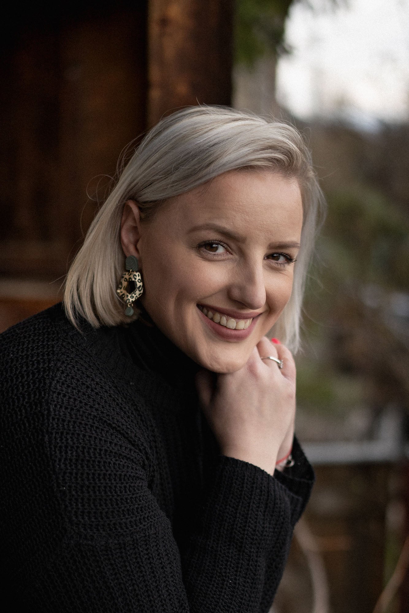 A woman with short blonde hair wearing a black sweater and striking Leopard Earrings smiles warmly while resting her chin on her hand. The background is softly blurred with natural, outdoor elements.