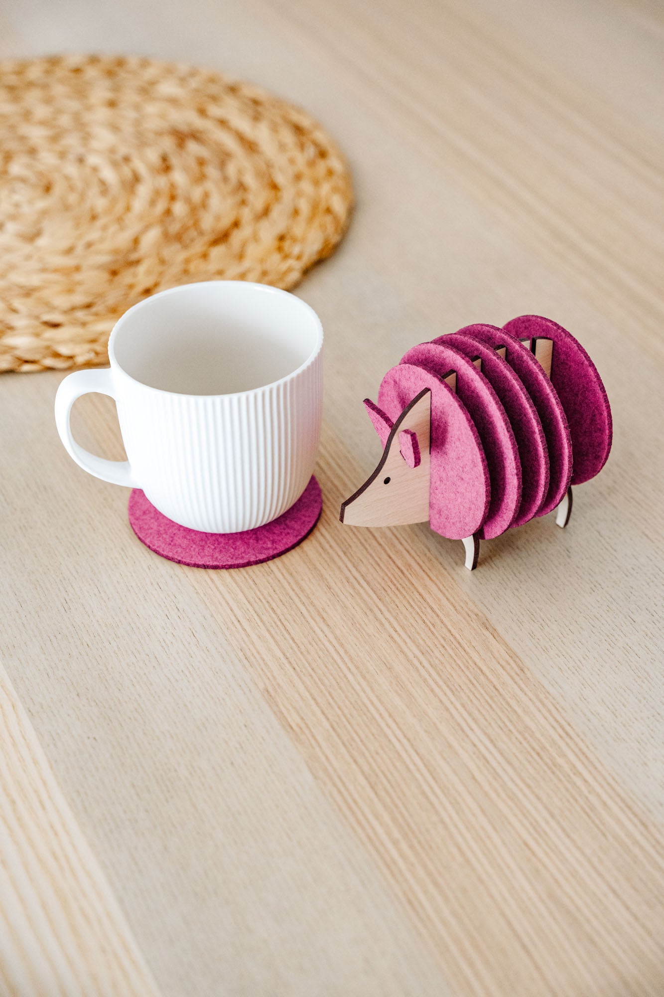 A white ribbed mug on a pink felt coaster sits next to a pink, hedgehog-shaped coaster holder on a light wooden table. A woven placemat is partially visible—perfect as a charming housewarming gift.