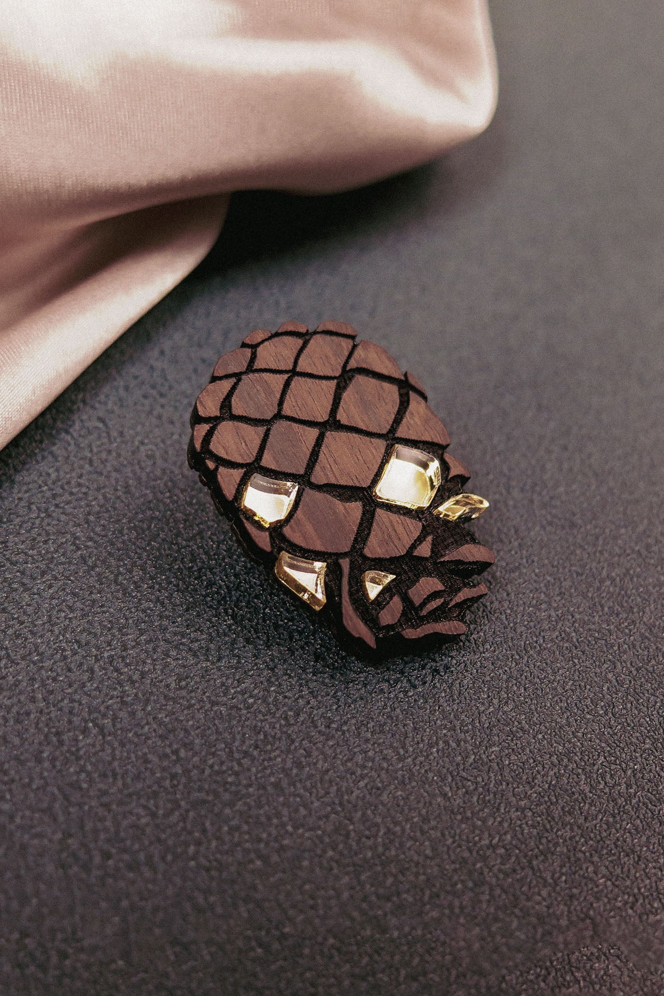 A sustainable pinecone brooch made from recycled wood with gold accents lies on a textured dark surface near a pink satin fabric.