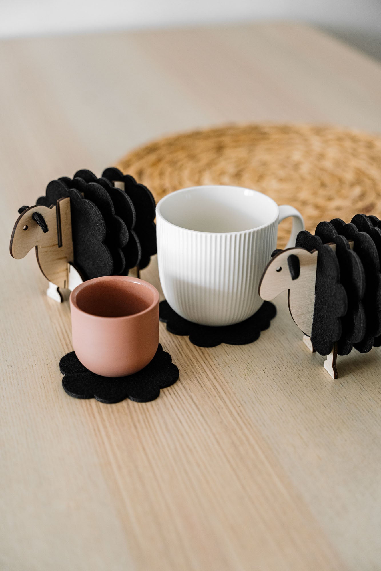 A white mug and a pink cup rest on felt coasters, accompanied by charming wooden sheep coaster holders on a light wooden table. Perfect as home decor accents, with a woven placemat in the background.