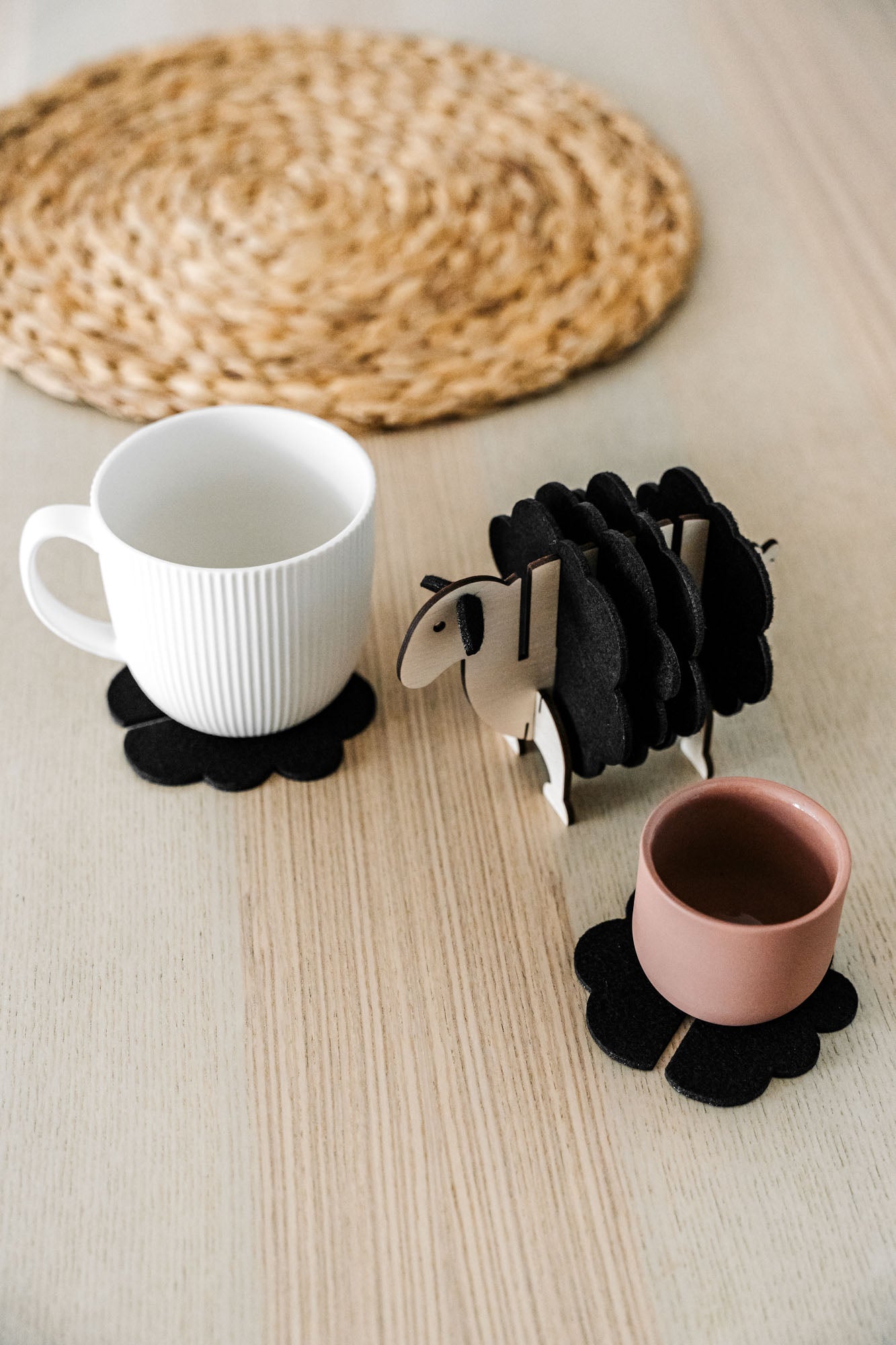 A white mug and a pink cup sit on black felt coasters next to a wooden sheep-shaped coaster holder on a light wooden table with a woven placemat in the background, perfect for charming home decor.