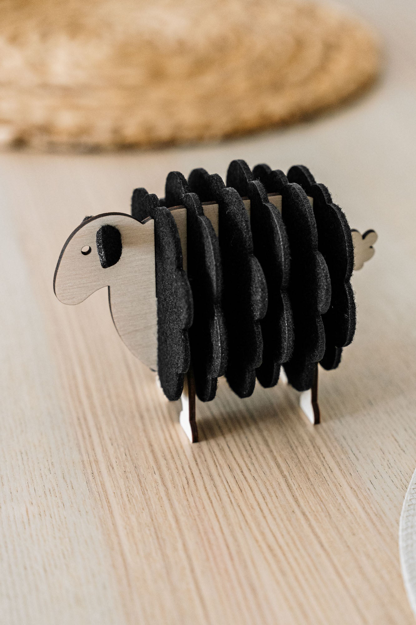 A charming wooden sheep-shaped coaster holder with black felt coasters stands on a light wooden table, bringing a touch of playful home decor alongside a woven placemat and white plate in the background.
