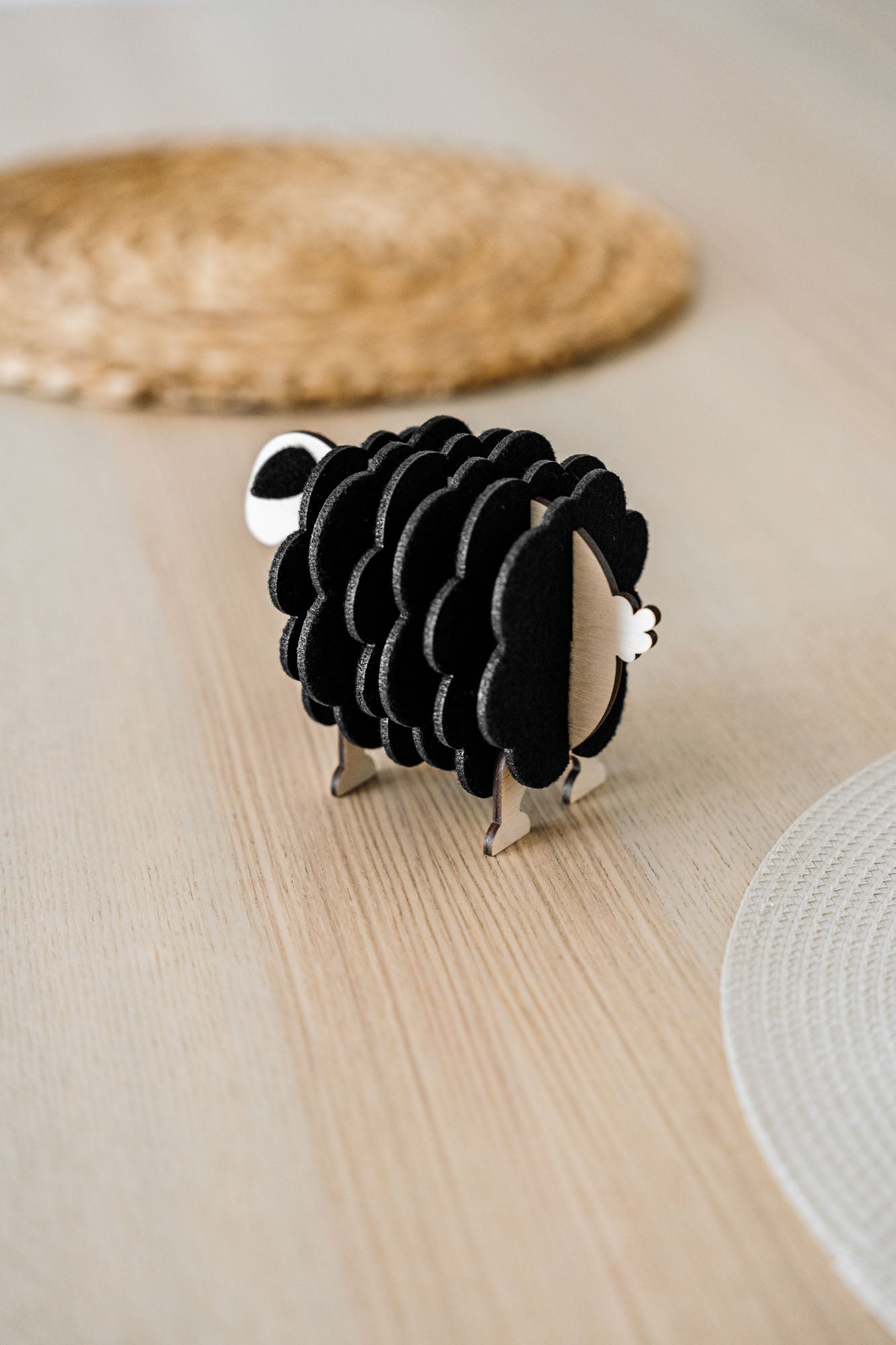 A small, black and white wooden sheep-shaped figure sits on a light wooden table near woven placemats, adding a charming touch to your home decor.