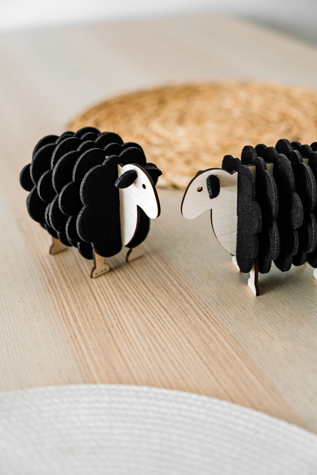 Two small, black wooden sheep figurines made of wood and felt stand facing each other on a light wooden surface, perfect for adding charm to your home decor, with a woven mat blurred in the background.