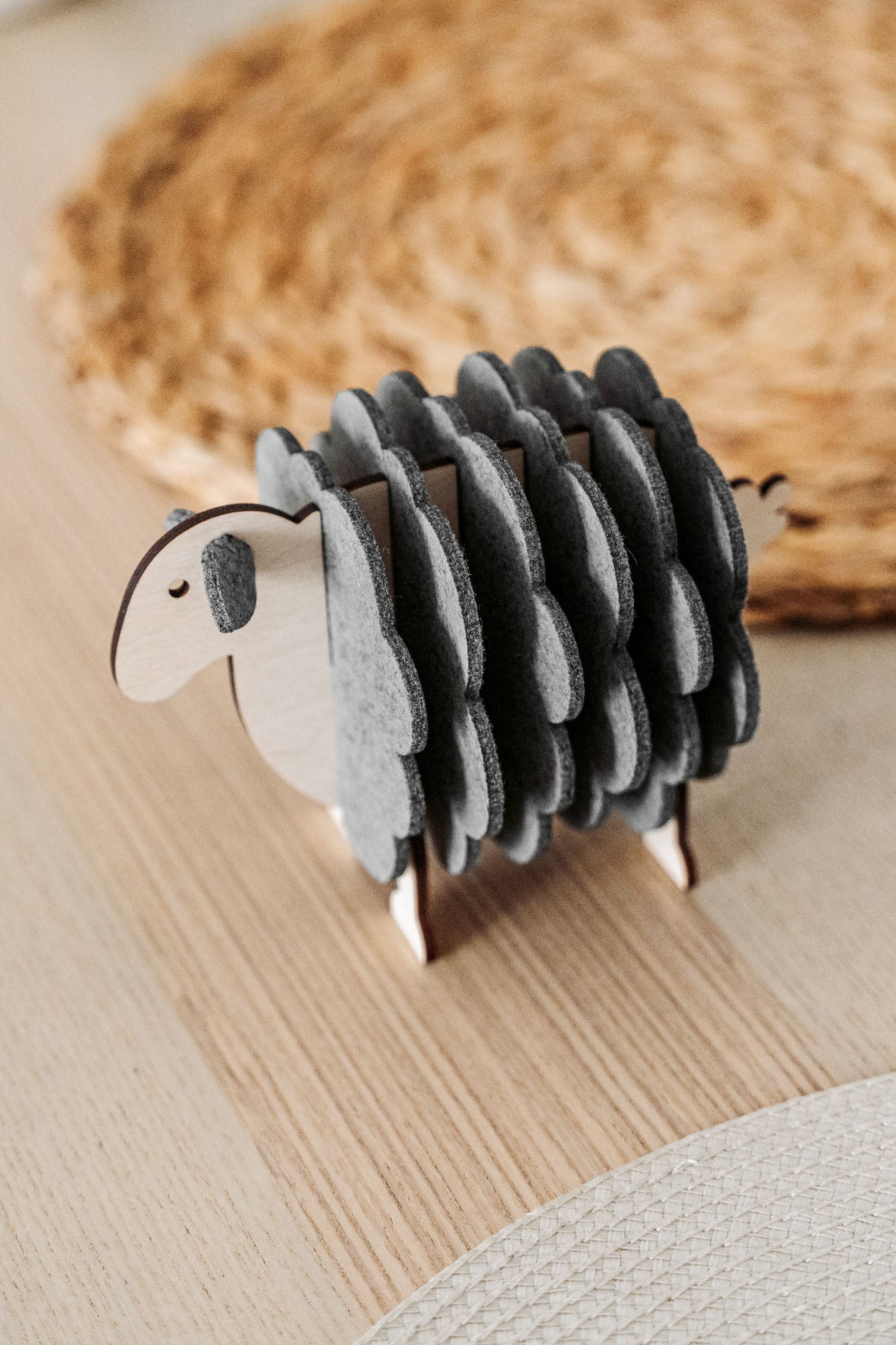 A small wooden sheep figurine with grey felt pieces for wool stands on a light wooden table, adding a charming touch of home decor, with a woven placemat in the background.