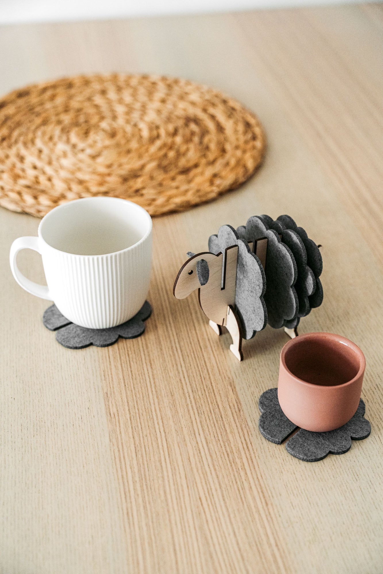 A white mug and a pink cup sit on dark felt coasters next to a decorative wooden sheep-shaped coaster holder, bringing charming home decor touches to a light wooden table topped with a round woven placemat.