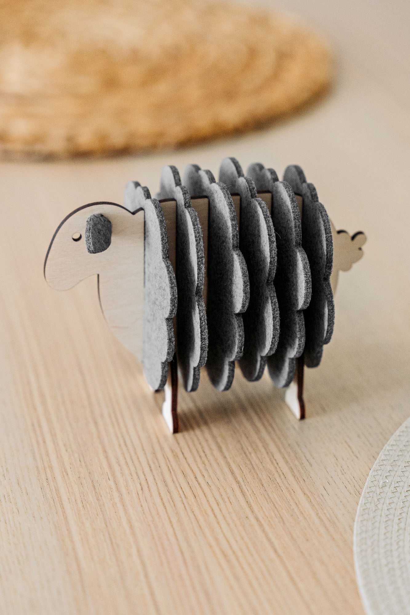 A wooden sheep-shaped coaster holder with round, gray felt coasters arranged to resemble the sheep’s wool stands on a light wooden table—perfect for charming home decor.