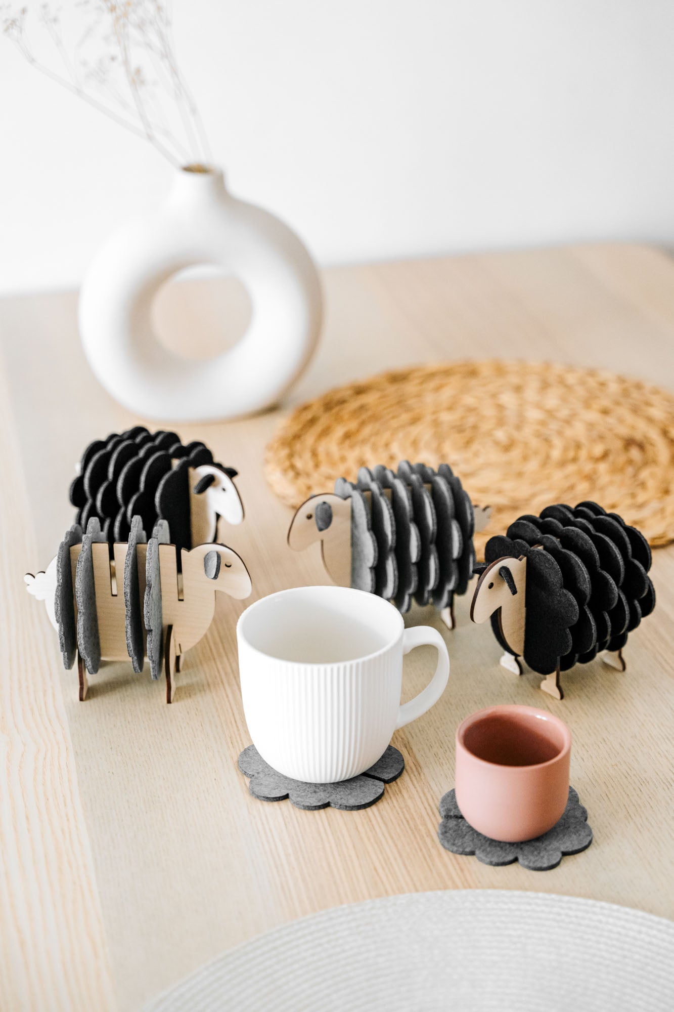 A white table holds a white vase, a woven placemat, and several sheep-shaped felt coasters. A white mug and a pink cup rest on two of the coasters, bringing playful charm to this cozy home decor setting.