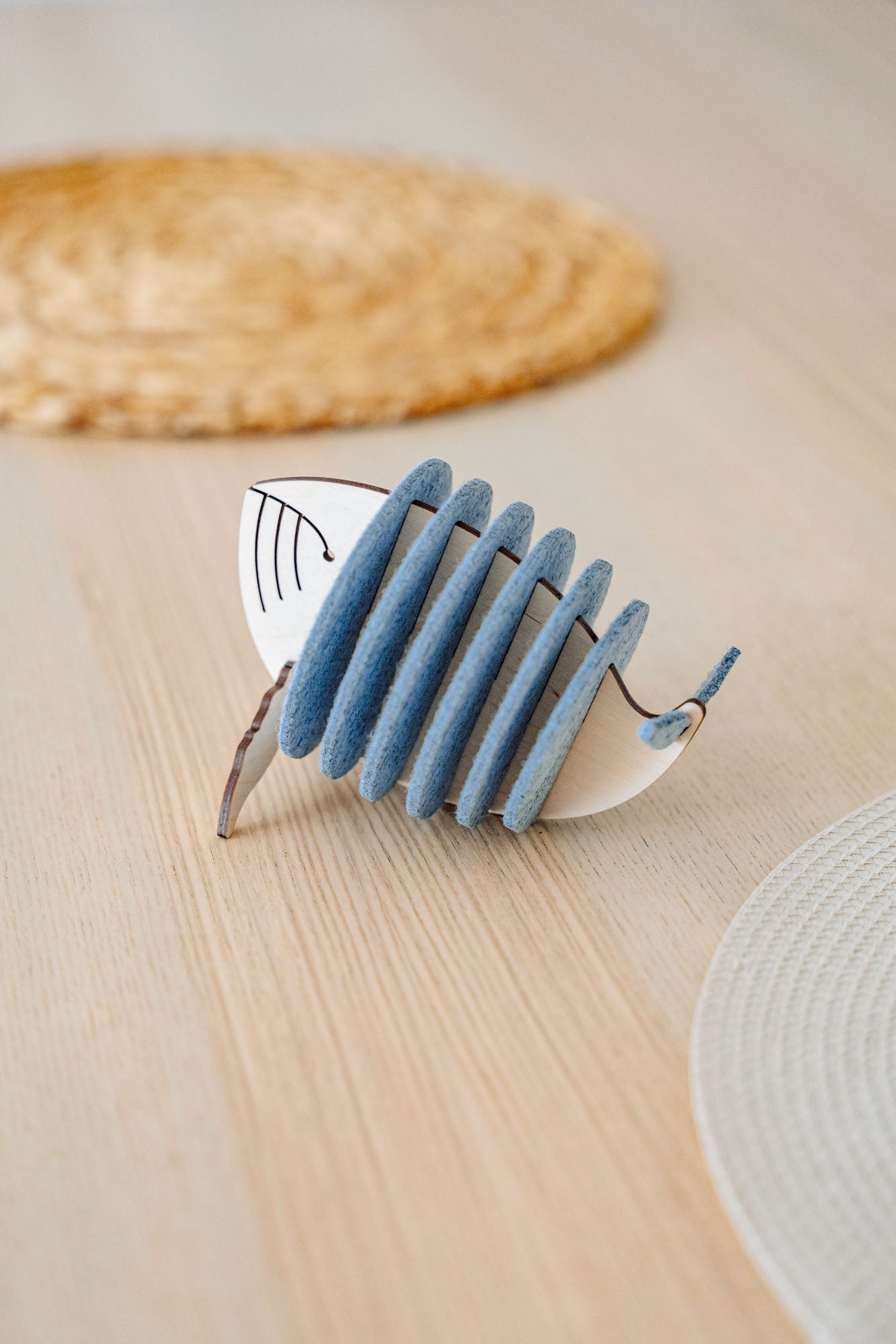 A decorative object resembling a stylized fish, made of alternating blue-grey felt and white pieces, sits on a light wooden table with woven placemats in the background, adding home charm and protecting surfaces like unique Whale Coasters.
