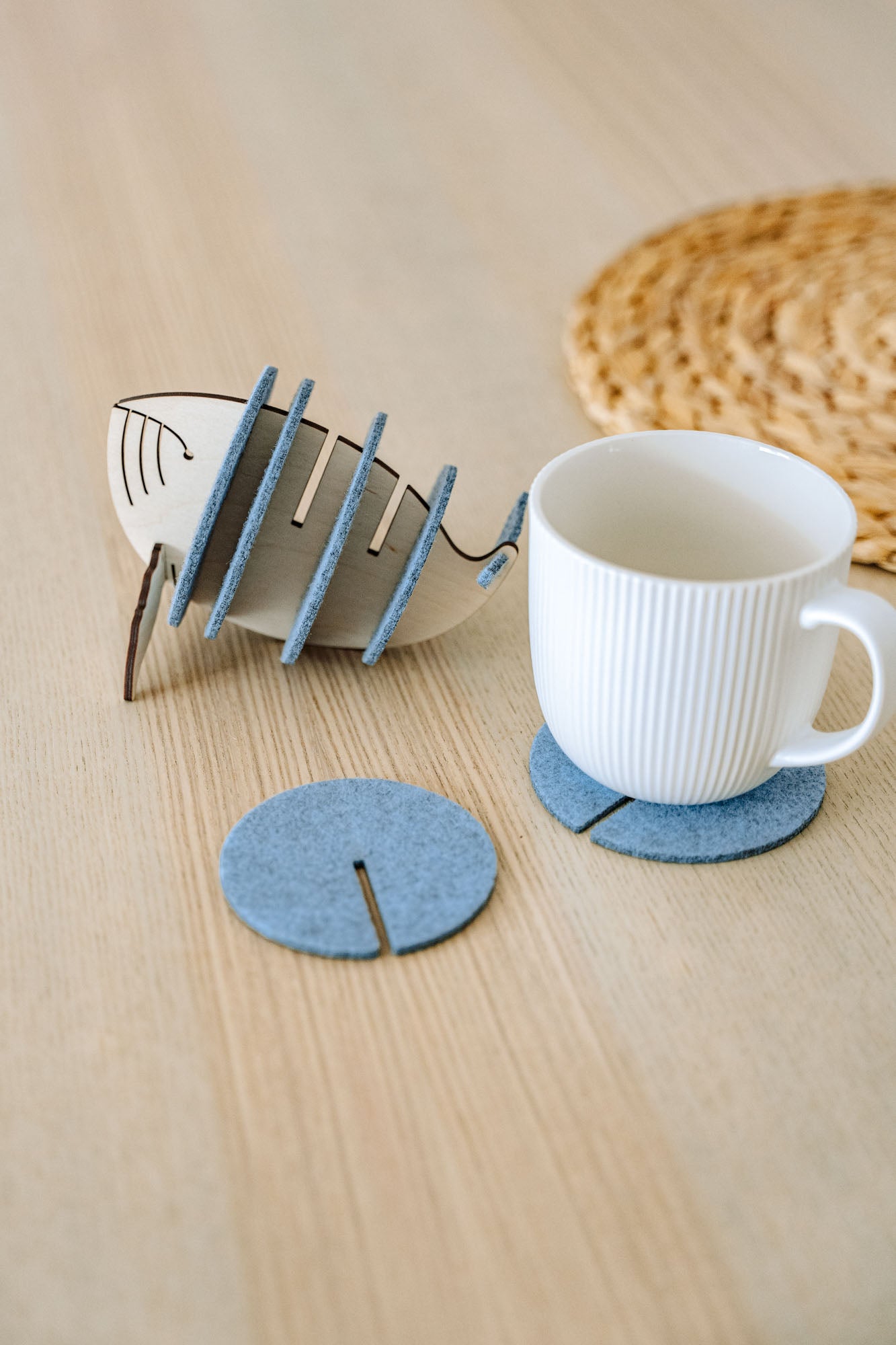 A white ribbed ceramic mug sits on a round gray felt coaster on a wooden table, with Whale Coasters and a whale-shaped holder nearby. A woven placemat adds home charm while these coasters help protect surfaces in style.