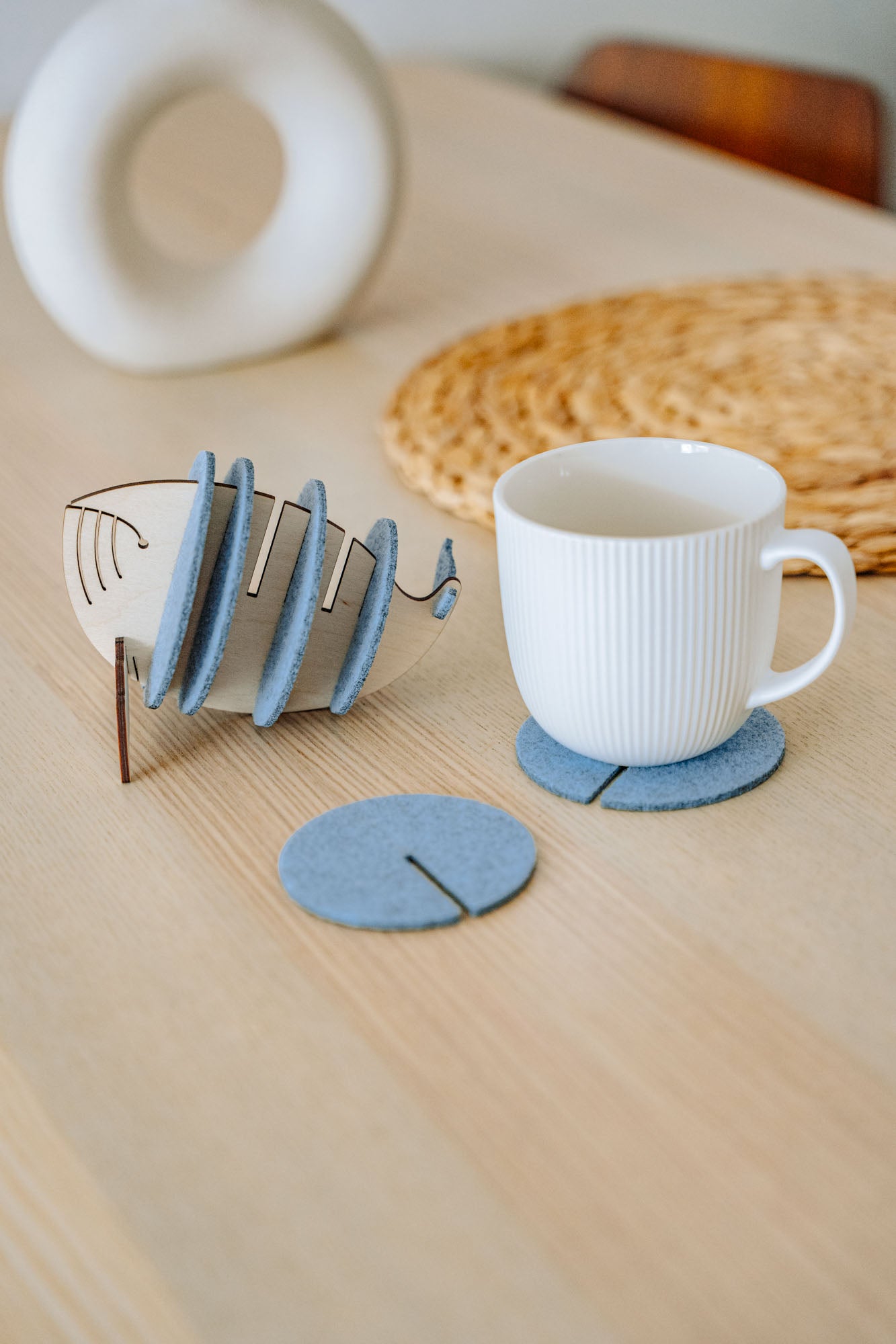 A white ribbed mug sits on a round gray felt coaster, designed to protect surfaces. Nearby, more Whale Coasters are stacked in a fish-shaped holder, with woven and ceramic decor adding home charm in the background.