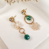 A pair of mismatched earrings displayed on a white surface, featuring green gemstones, gold accents, and white floral-shaped elements, exuding timeless elegance with delicate dried grass in the background.
