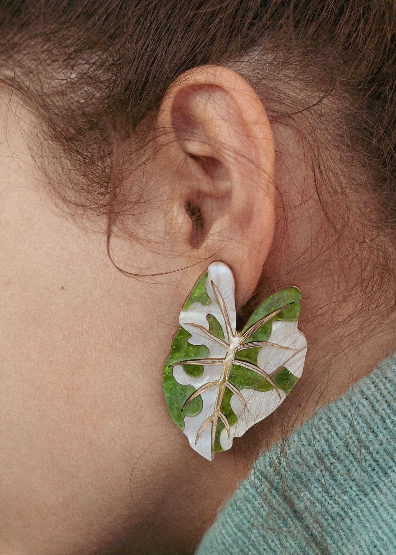A close-up of a persons ear adorned with large, leaf-shaped Alocasia Earrings in green and white. The hand-crafted jewelry stands out against their brown hair and a light blue-green garment.