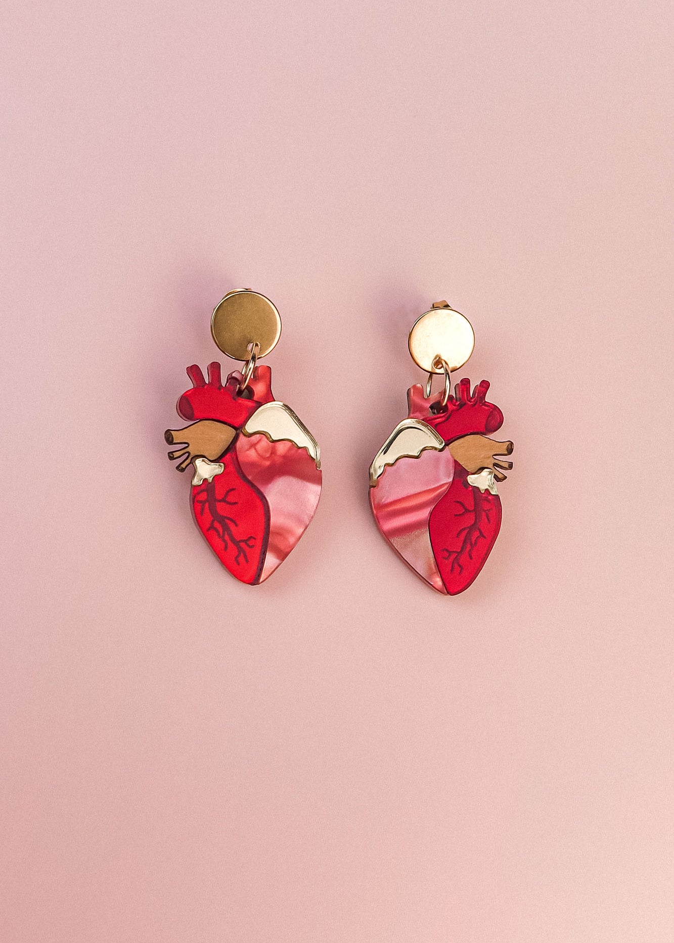 A pair of gold and pink Anatomical Heart Earrings, crafted in acrylic with detailed vein patterns and a unique style, displayed on a light pink background.
