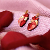 Two red Anatomical Heart Earrings with gold accents rest on a soft, pink textured fabric, surrounded by red rose petals—a unique style statement for acrylic earrings lovers.