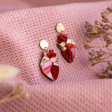 A pair of red and gold Anatomical Heart Earrings rest on textured pink fabric, with dried flowers nearby—acrylic earrings perfect for expressing your unique style.