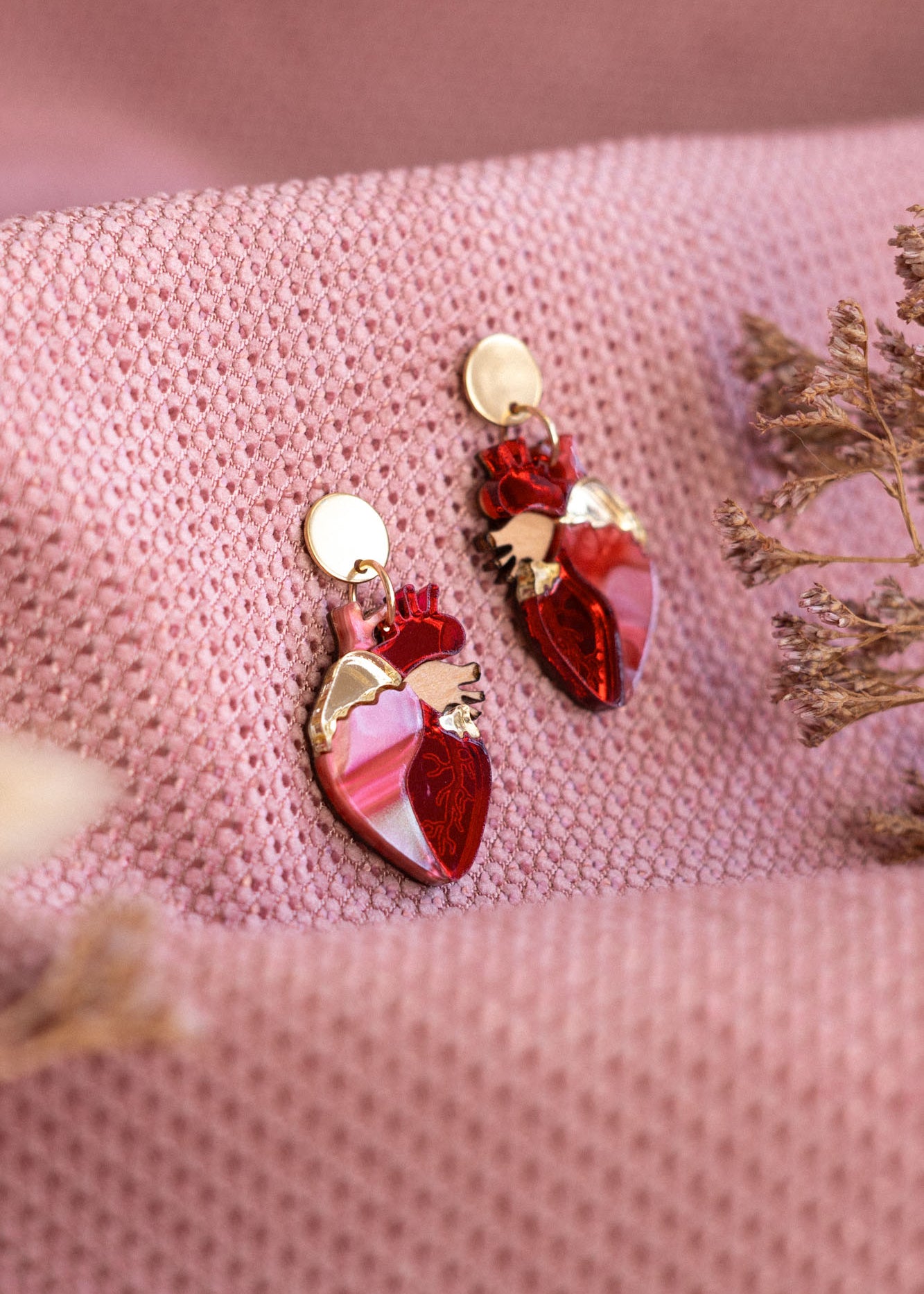 A pair of red and gold Anatomical Heart Earrings rest on textured pink fabric, with dried flowers nearby—acrylic earrings perfect for expressing your unique style.