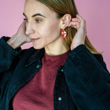 A woman with light brown hair wears a maroon shirt and black jacket, adjusting her unique acrylic earrings. She stands in front of a pink background, looking to the side with a slight smile.