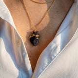 A person wearing a white button-up shirt with two layered gold necklaces, including a unique anatomical heart necklace. The scene is lit by warm sunlight.