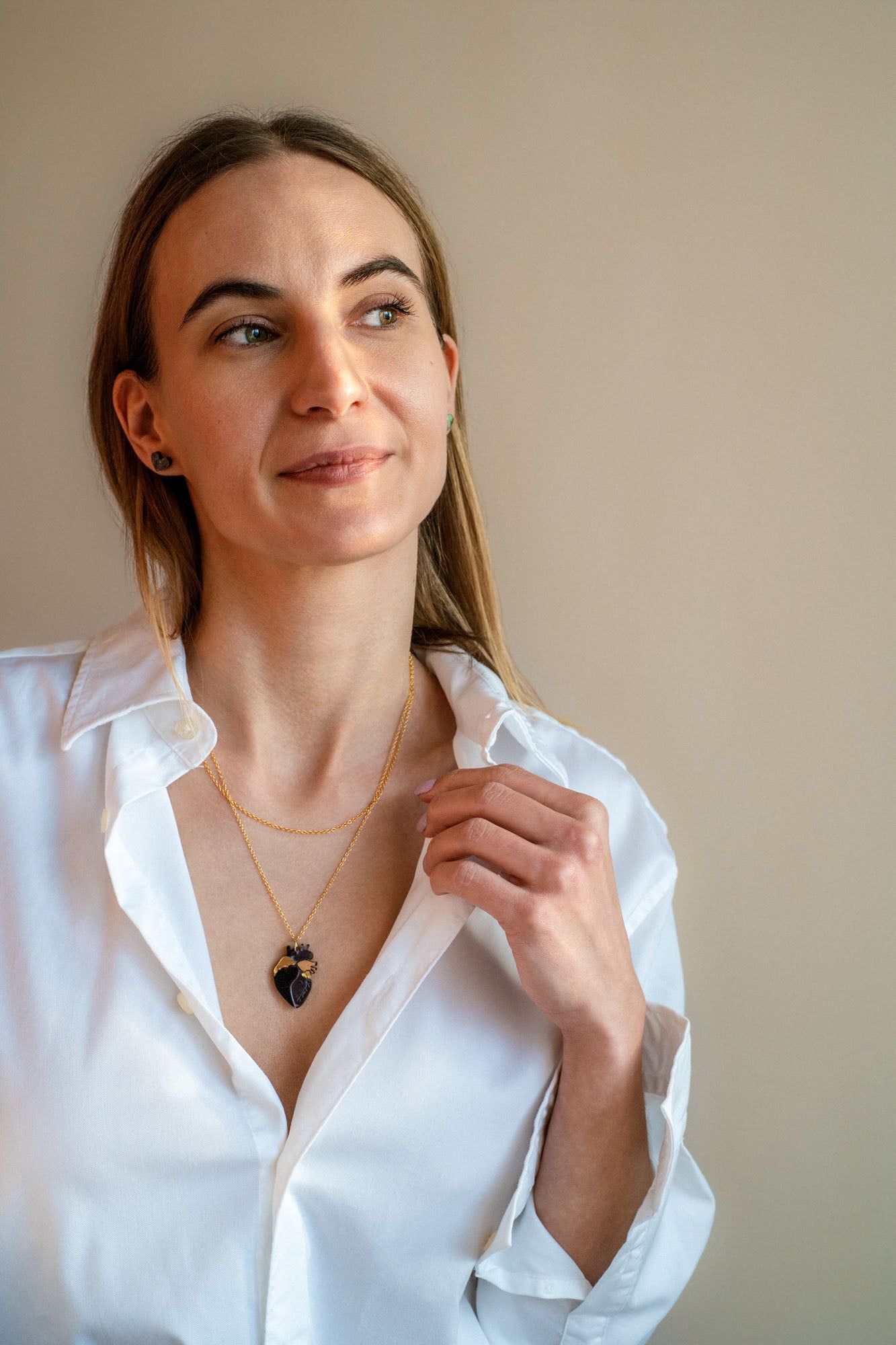 A woman with long light brown hair wears a white button-up shirt and a gold anatomical heart necklace, looking slightly to the side with a soft smile against a plain beige background.