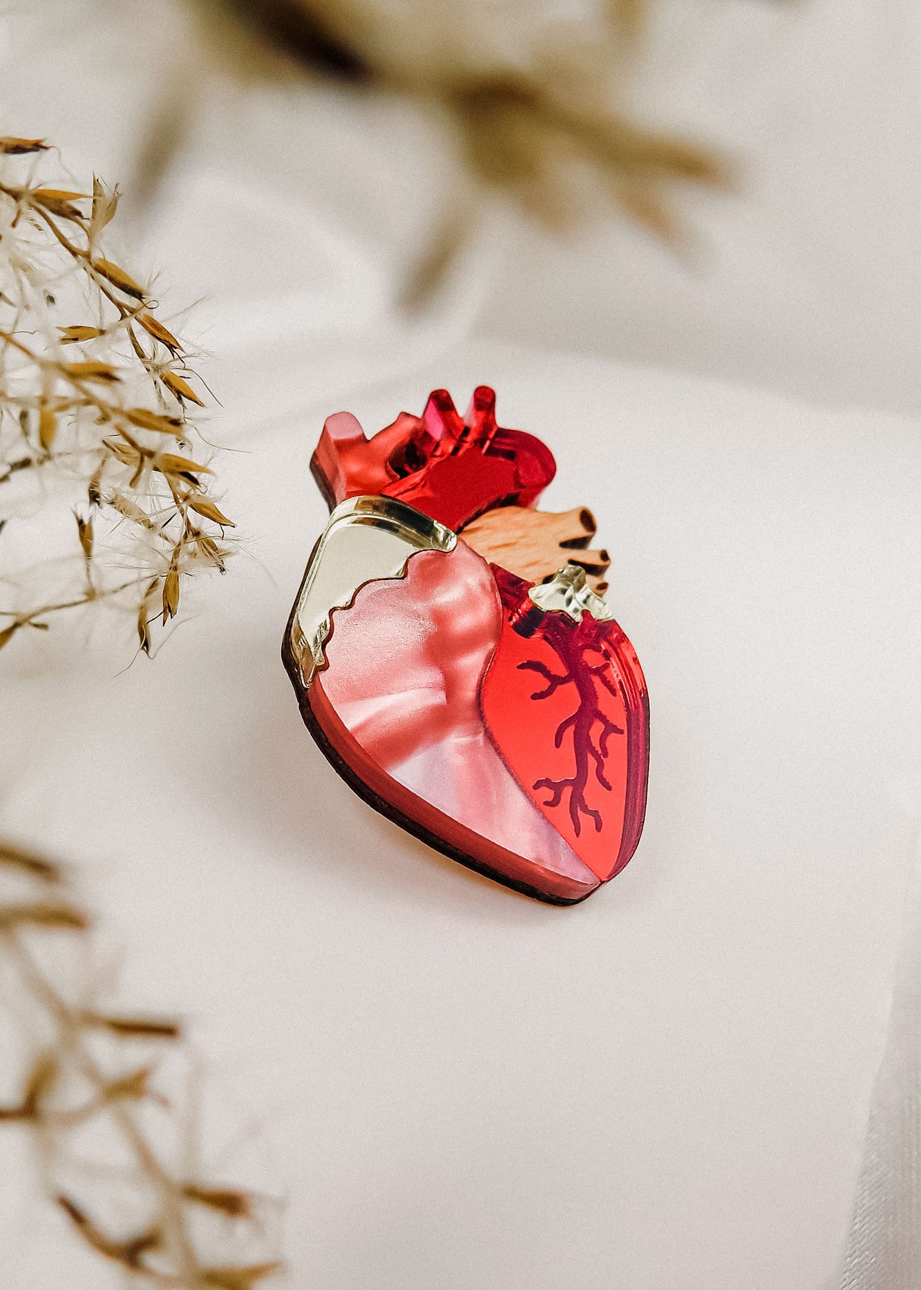A handcrafted perfume bottle shaped like an anatomical heart, featuring red and pink tones with vein-like details, rests on a soft white surface beside dried grasses—truly a statement piece for any collection.