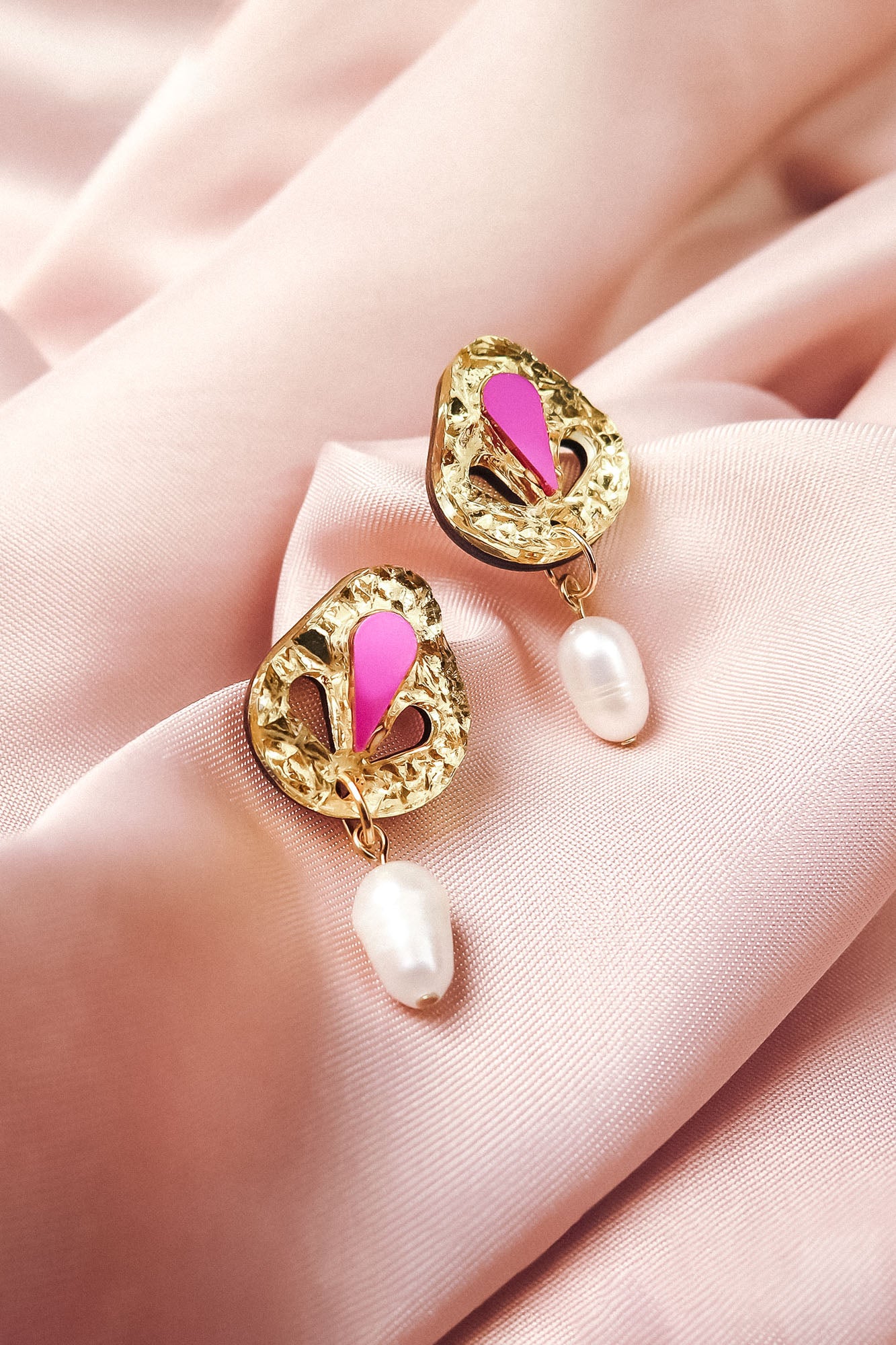 A pair of handcrafted gold drop earrings with intricate designs, pink enamel accents, and white pearl drops, beautifully displayed on soft, pale pink fabric.