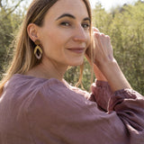 A woman with long light brown hair sits outdoors, smiling slightly as she looks over her shoulder. She wears a mauve blouse and handcrafted Bear Earrings made from premium acrylic glass, with sunlight and greenery in the background.