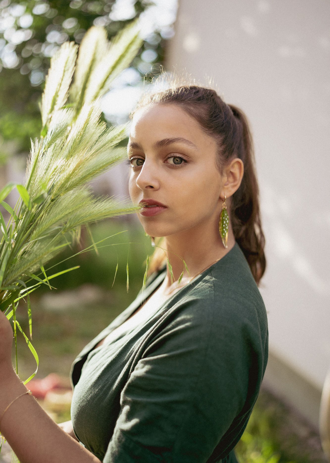 A woman with long brown hair in a ponytail, wearing a dark green top and unique jewelry—Begonia Leaves Earrings—stands outdoors in sunlight holding tall grass, looking directly at the camera. The softly blurred background is perfect for plant lovers.