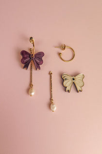 Three mismatched earrings on a pink background: a gold-plated hoop earring, a purple bow with a dangling freshwater pearl, and a white bow edged in gold attached to a bar and pearl—perfect for creating your own interchangeable dangle earring look.