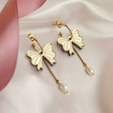 A pair of gold-plated earrings shaped like bows with dangling freshwater pearl accents, displayed on white satin fabric with a pink ribbon and gold decorative balls nearby.
