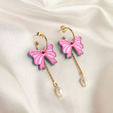 A pair of gold-plated c-hoop earrings with pink butterfly charms and freshwater pearl drops, displayed on white satin fabric with decorative gold spheres nearby.