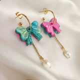 Gold-plated c-hoop earrings with colorful butterfly charms—one blue and one pink—feature freshwater pearl and gold chain accents. These interchangeable dangle earrings are displayed on a white fabric background.
