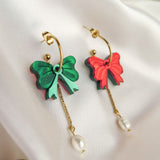 Two gold-plated c-hoop earrings with bow-shaped charms, one green and one red, each featuring a freshwater pearl dangling from a gold chain—stylish interchangeable dangle earrings displayed on white fabric.