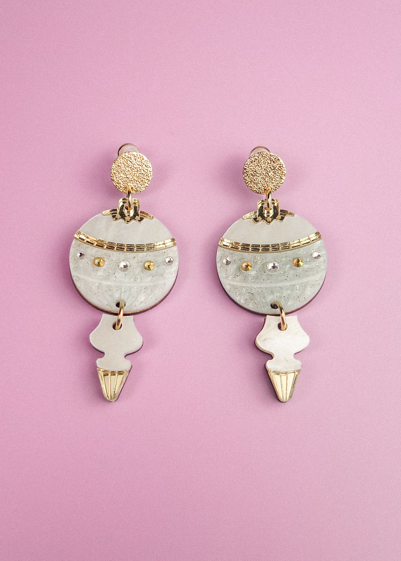 A pair of ornate, gold and white festive earrings with circular and abstract shapes, resting on a pastel purple background.