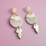 A pair of decorative dangle earrings with gold and silver accents, featuring round and arrow-shaped elements, make for stunning festive earrings, beautifully displayed on a light purple background.