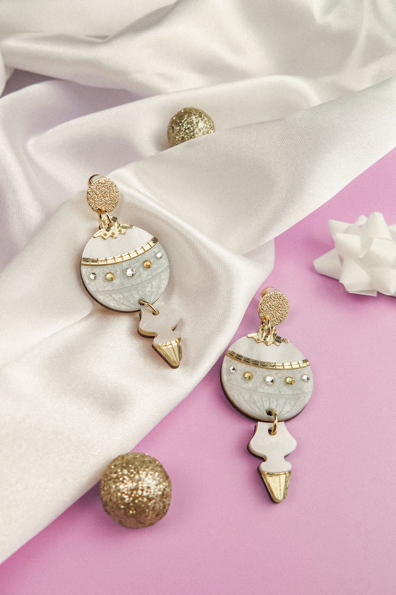 A pair of gold and white festive earrings with pearl details rest on white satin fabric, surrounded by a glittery gold ball and a white decorative bow on a lavender background.