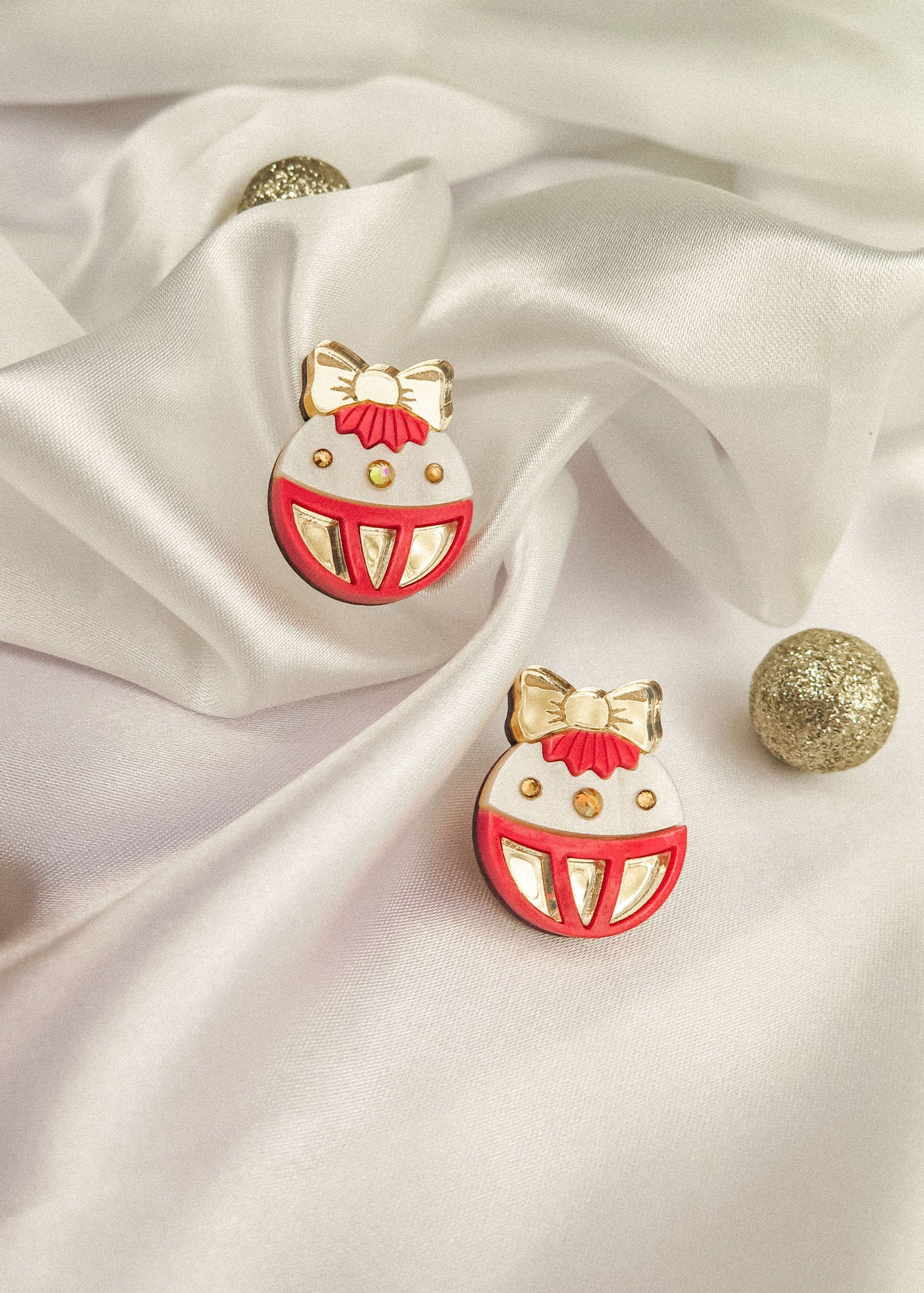 A pair of round enamel pins with red, white, and gold details, resembling festive ornaments with bows, are displayed on white satin fabric with gold glittery balls—perfect as holiday jewelry or a festive earrings-inspired accent.